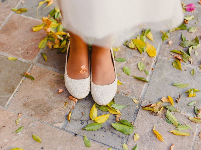 Eine Person in cremefarbenen flachen Schuhen steht auf einem mit gelben und grünen Blättern übersäten Fliesenboden. Auf einem Schuh ist ein kleiner dekorativer Schmetterling.