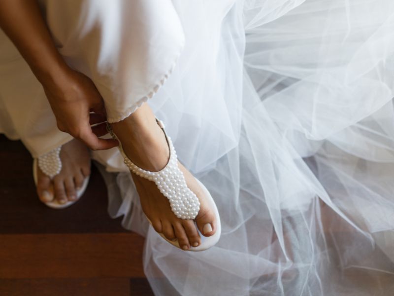 Eine Person im Brautkleid rückt eine mit Perlen besetzte Sandale an ihrem Fuß zurecht.