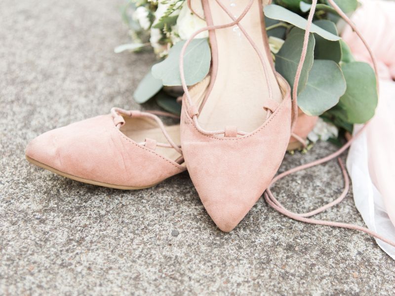 Ein Paar hellrosa Ballerinas mit langen Schnürriemen auf einer Betonoberfläche mit einigen grünen Blättern und einem Stück weißem Stoff im Hintergrund.