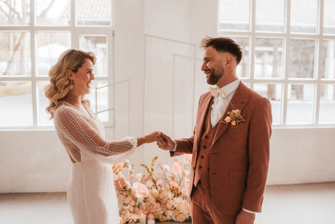 Ein Paar im Hochzeitskleid hält Händchen und lächelt sich in einem hellen Raum mit großen Fenstern und Blumenarrangements an.