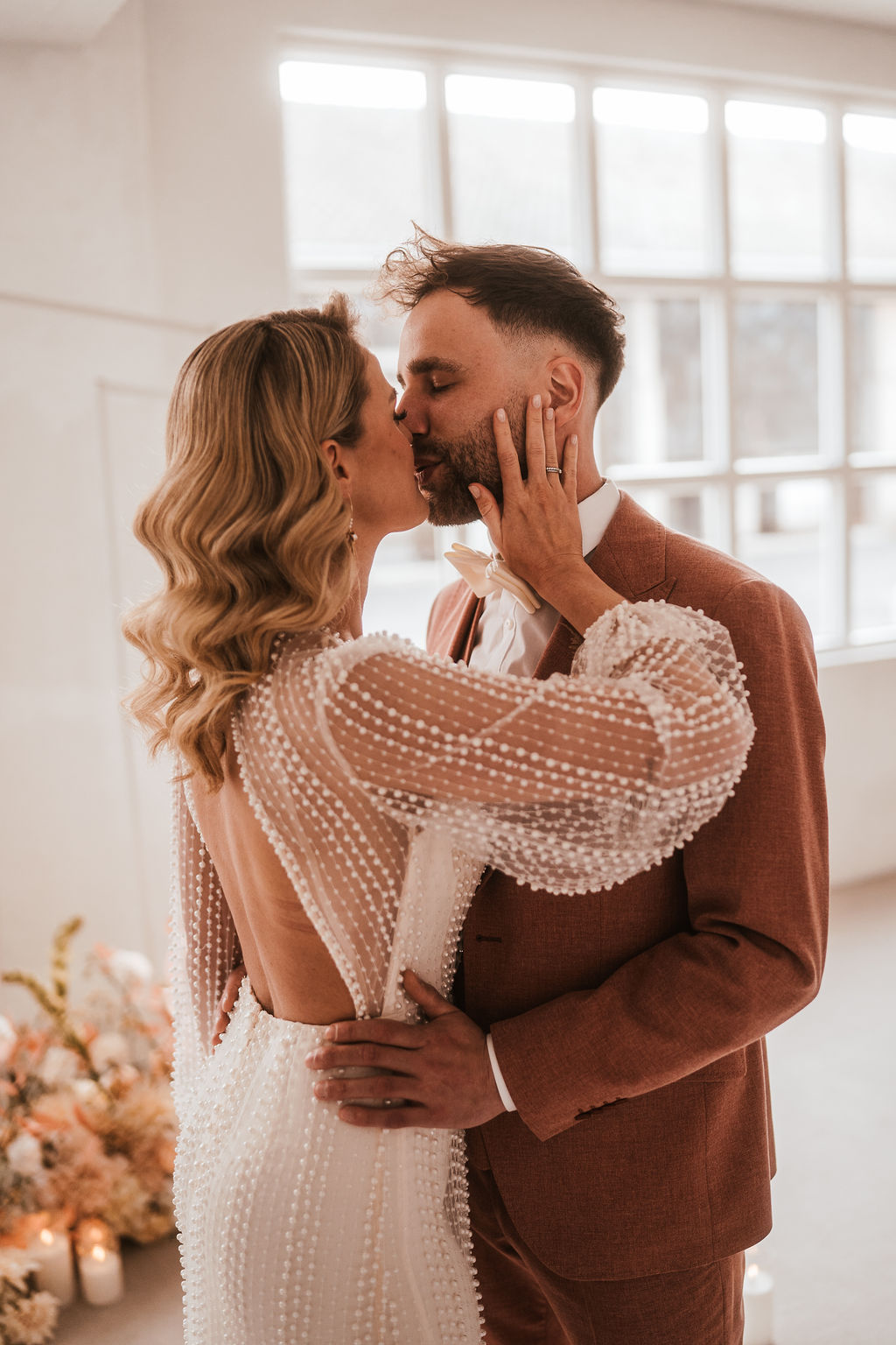 Ein Paar küsst sich drinnen. Die Frau trägt ein weißes Kleid mit perlenartigen Verzierungen und der Mann einen braunen Anzug mit weißem Hemd. Im Hintergrund sind sanfte Beleuchtung und Blumenarrangements zu sehen.