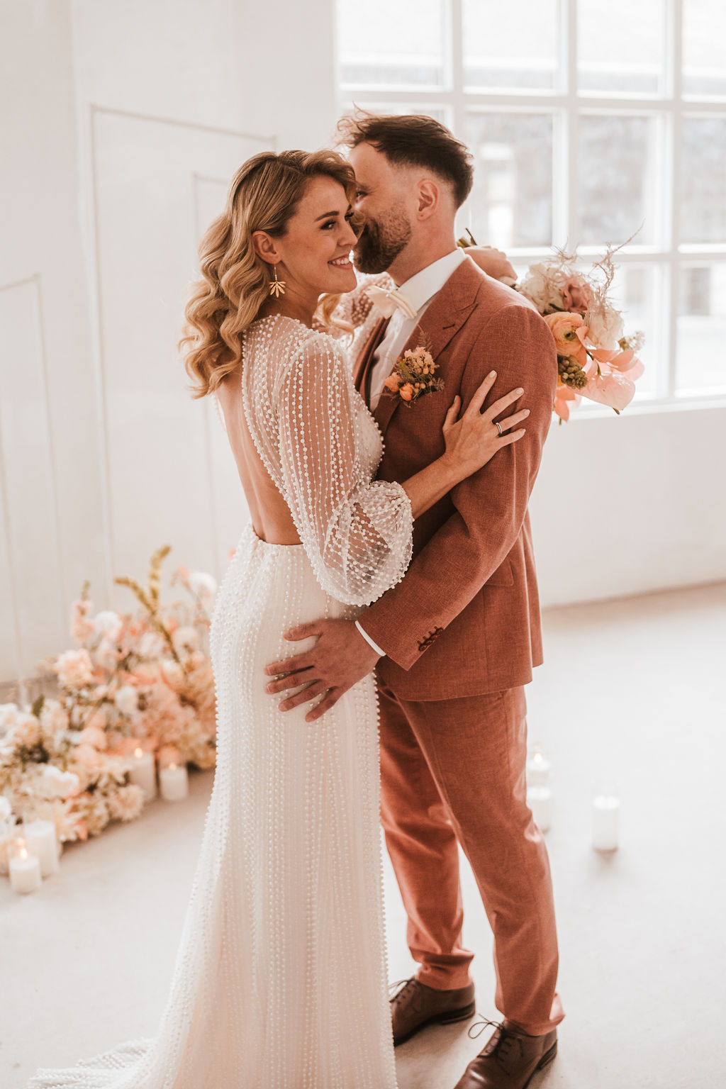 Ein Paar im Hochzeitskleid steht dicht beieinander in einem gut beleuchteten Raum mit Blumenschmuck und Kerzen. Die Frau trägt ein langes weißes Kleid und der Mann einen orangefarbenen Anzug mit einem Blumenstrauß hinter ihnen.