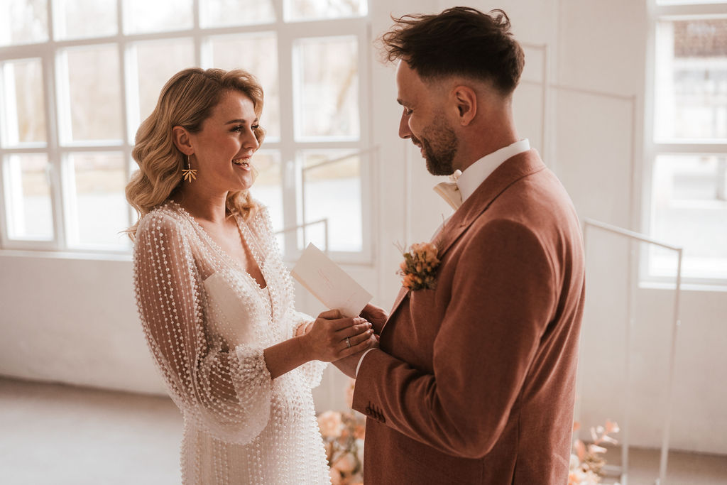 Braut und Bräutigam geben sich drinnen das Ja-Wort. Sie trägt ein weißes, mit Perlen besetztes Kleid und hält eine Karte in der Hand, während er einen hellbraunen Anzug und eine Ansteckblume trägt. Beide lächeln sich an.