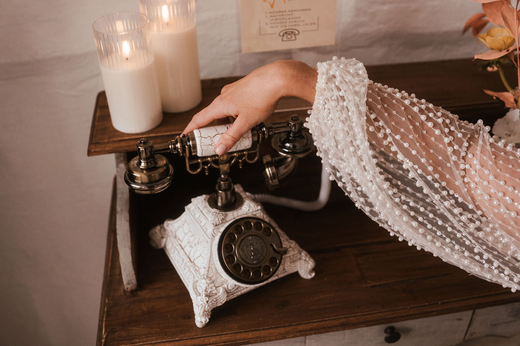 Eine Hand in einem Spitzenärmel greift nach dem Hörer eines reich verzierten Vintage-Telefons mit Wählscheibe auf einem Holztisch mit brennenden Kerzen im Hintergrund.