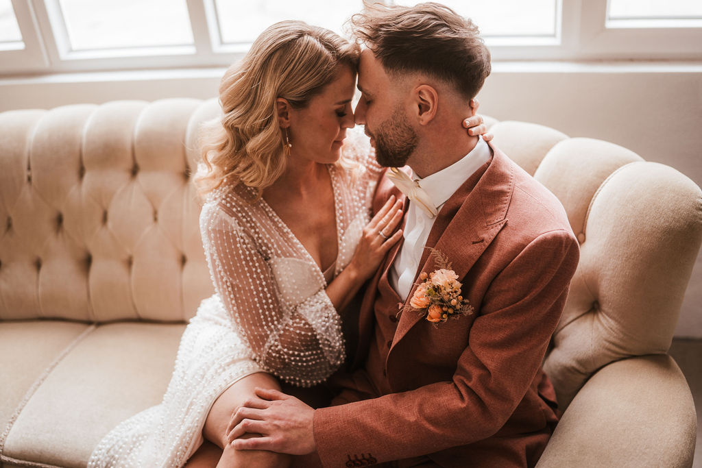 Ein Paar sitzt auf einem cremefarbenen Sofa und teilt einen zärtlichen Moment, wobei sich ihre Stirnen berühren. Die Frau trägt ein weißes Kleid und der Mann einen rosa Anzug mit einer Ansteckblume.