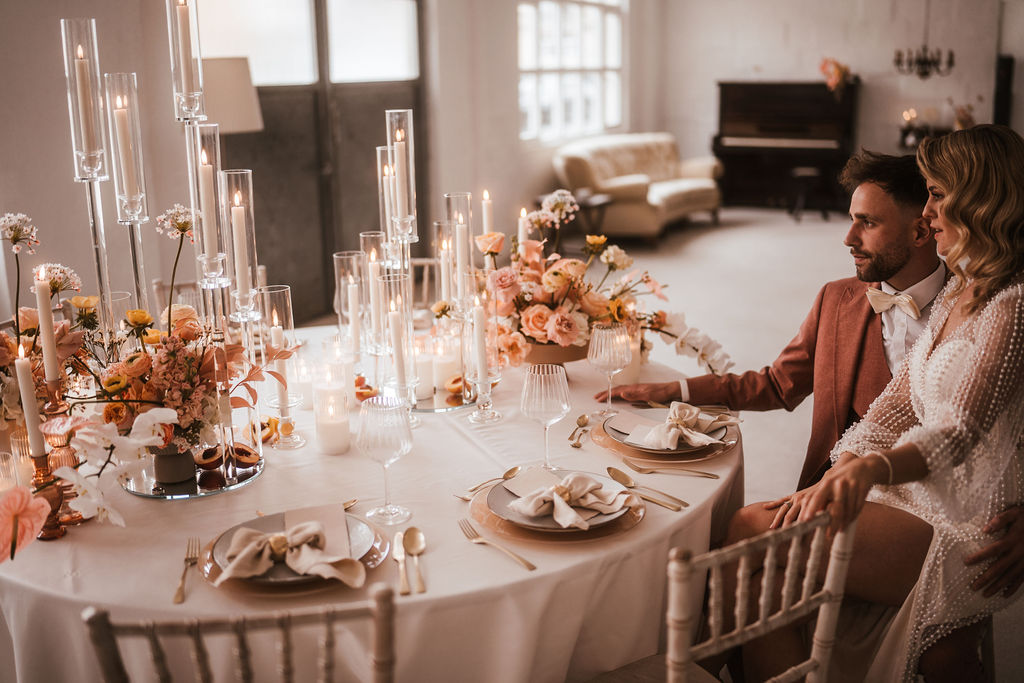 Ein Paar sitzt an einem wunderschön dekorierten Tisch mit Blumenarrangements, brennenden Kerzen und Gedecken in einem warm beleuchteten Raum mit einem Klavier im Hintergrund.