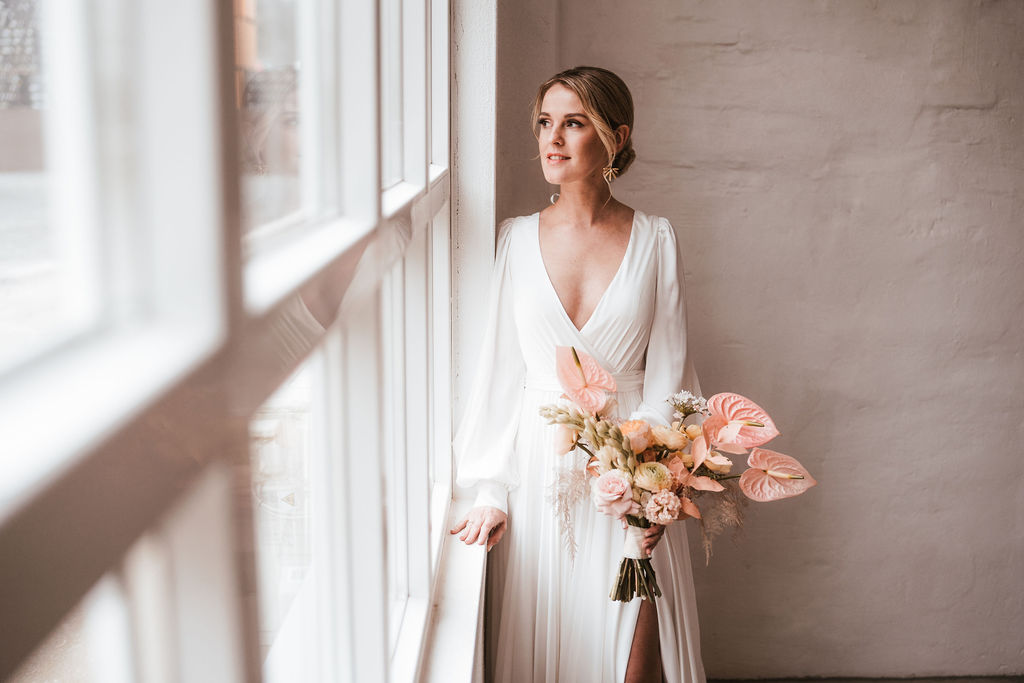 Eine Braut in einem weißen Kleid hält einen Strauß rosa und cremefarbener Blumen in der Hand und steht an einem Fenster und schaut nach draußen.