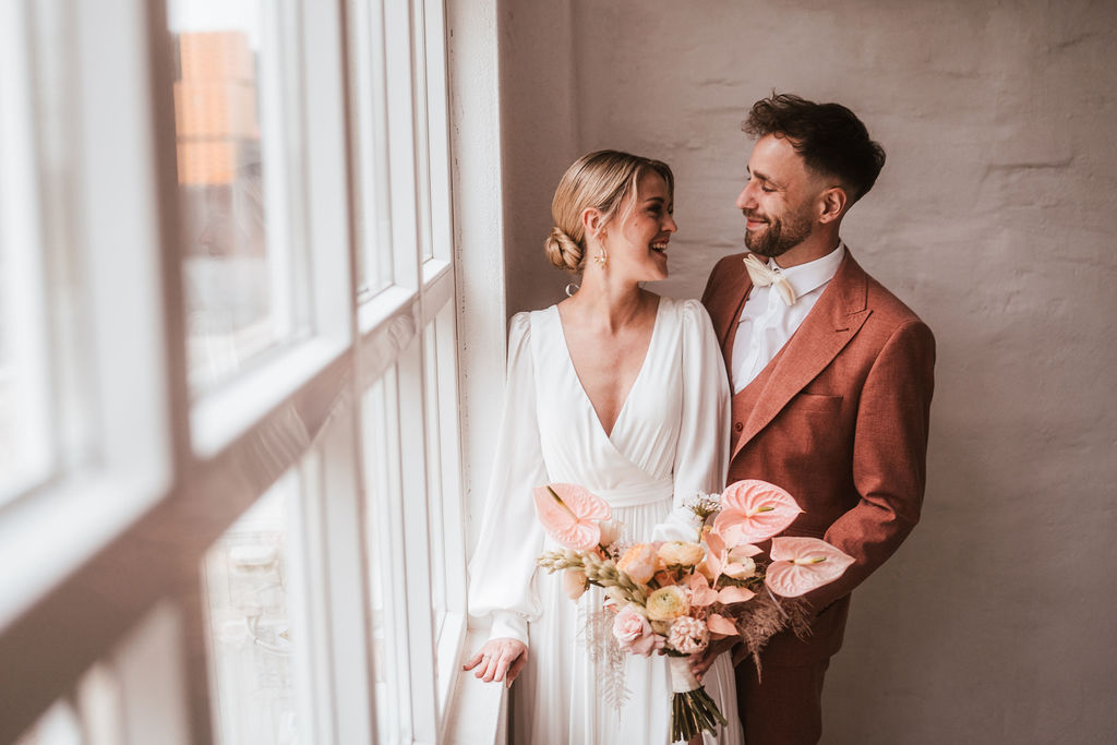 Ein Paar im Hochzeitskleid steht an einem Fenster und lächelt sich an. Die Braut hält einen Blumenstrauß.