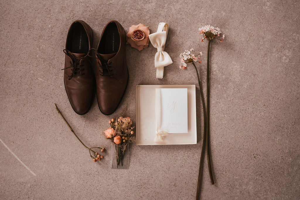 Flache Anordnung aus braunen Abendschuhen, einer Boutonniere, Blumen, einer Fliege, einer Rose und einer weißen Einladungskarte mit Schleife auf einer beigen Oberfläche.