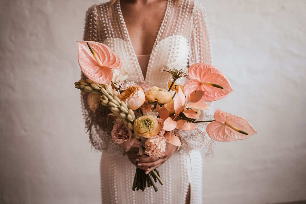 Eine Person in einem weißen, perlenbesetzten Kleid hält einen Strauß orangefarbener und rosa Blumen vor einem schlichten Hintergrund.