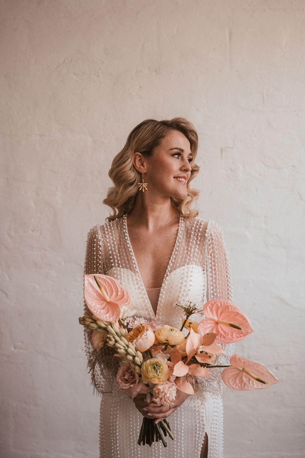 Eine Frau in einem weißen Kleid hält einen Strauß rosa und weißer Blumen und blickt vor einem schlichten Hintergrund nach links.